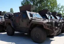 Line of armored military vehicles parked outdoors, tan colored, with angled armor and mounted equipment on top.