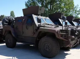 Line of armored military vehicles parked outdoors, tan colored, with angled armor and mounted equipment on top.