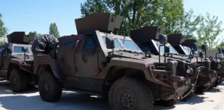 Line of armored military vehicles parked outdoors, tan colored, with angled armor and mounted equipment on top.