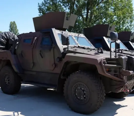 Line of armored military vehicles parked outdoors, tan colored, with angled armor and mounted equipment on top.