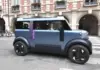 Cred că Brevetarea DACIA Hipster este o Greșeală a DACIA Blue futuristic car with a purple stripe drives along a city street in front of a brick building with arches and balconies.