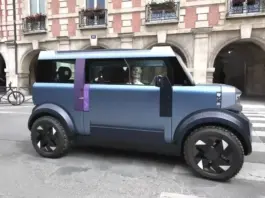 Blue futuristic car with a purple stripe drives along a city street in front of a brick building with arches and balconies.