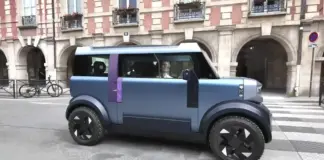 Blue futuristic car with a purple stripe drives along a city street in front of a brick building with arches and balconies.
