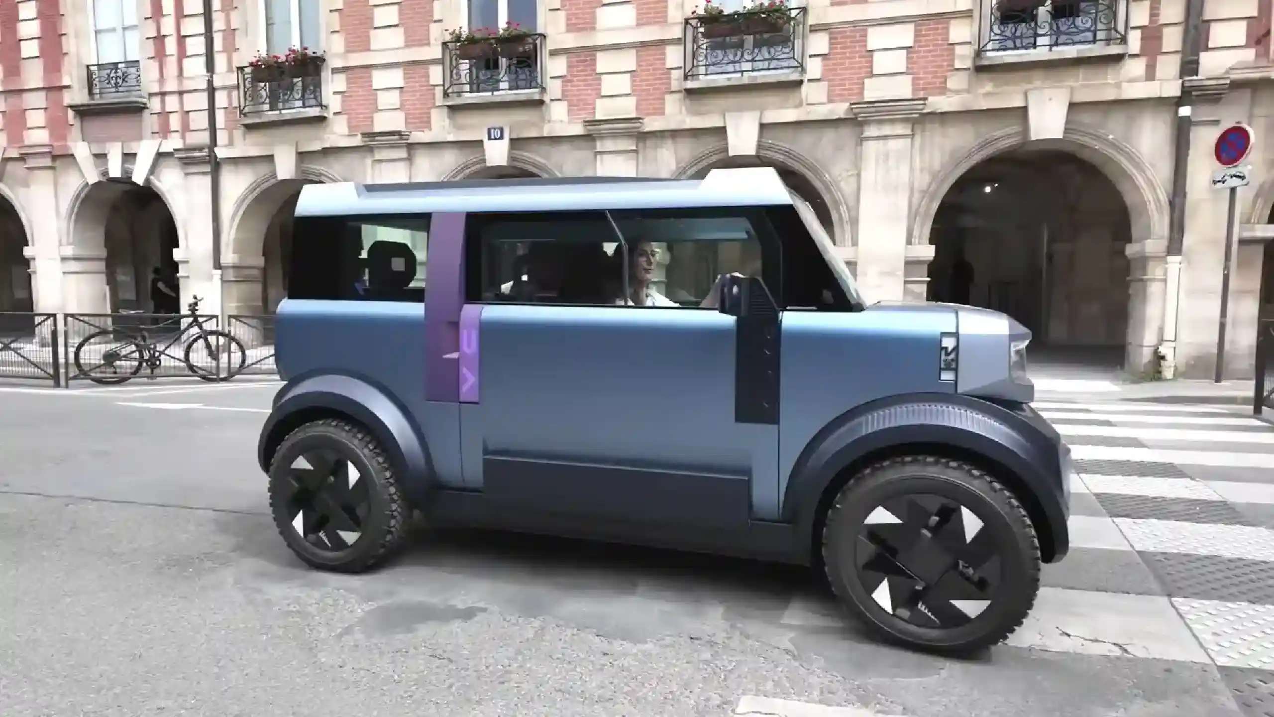 DACIA Hipster brevet | iDevice.ro Blue futuristic car with a purple stripe drives along a city street in front of a brick building with arches and balconies.