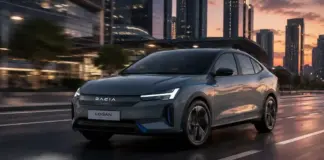 Gray Dacia Logan Electric car driving on a city street at sunset with tall buildings in the background.