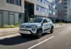 Silver compact SUV driving along a city street with modern glass-fronted buildings on both sides, motion blurred lanes.