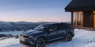 Grey SUV parked on a snow-covered driveway beside a wooden cabin, with mountains in the distance at dusk.