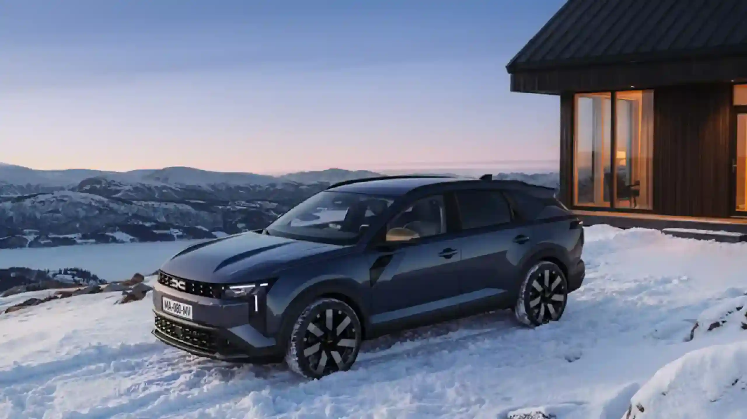 DACIA Striker 2026 lansare | iDevice.ro Grey SUV parked on a snow-covered driveway beside a wooden cabin, with mountains in the distance at dusk.