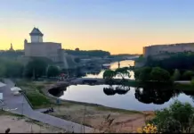 Decizia RADICALA a Estoniei din Cauza Amenintarii Rusiei Fortified castle walls and towers along a calm river at sunset, with greenery in the foreground and a pedestrian pathway nearby.