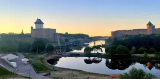 Fortified castle walls and towers along a calm river at sunset, with greenery in the foreground and a pedestrian pathway nearby.