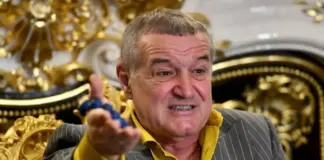 Man in a pinstripe suit and yellow shirt gesturing as he speaks in a lavish, gold-adorned room.