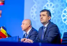 Ministrul Apararii ATENTIONEAZA Romanii dupa Atacul Navei din Marea Neagra Two suited men sit at a conference panel on a blue stage, a Romanian flag visible on the desk in front of them.