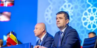 Two suited men sit at a conference panel on a blue stage, a Romanian flag visible on the desk in front of them.
