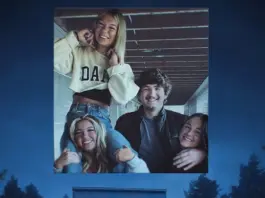 Group of five friends smiling and posing together in a large photo mounted outdoors, with arms around each other in a friendly stack at dusk/sunset.