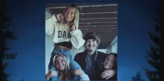 Group of five friends smiling and posing together in a large photo mounted outdoors, with arms around each other in a friendly stack at dusk/sunset.