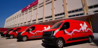 Several red delivery vans with green and white decorative graphics parked in front of a beige multi-story building under a blue sky, with a pink banner on the facade