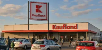 Exterior view of a Kaufland supermarket with a large red sign and logo, and a crowded parking lot in front.