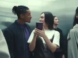 Two friends share a playful moment in a crowded elevator, one woman showing a phone to the other as they smile at each other.