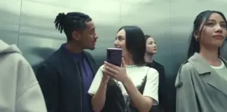 Two friends share a playful moment in a crowded elevator, one woman showing a phone to the other as they smile at each other.