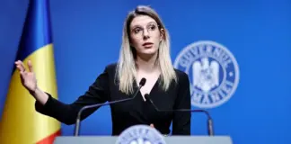 Woman speaking at a podium with a raised hand, Romanian flag on left and blue backdrop with official seal behind her.