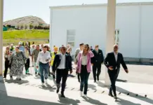 Premiera Anuntata de Ministrul Mediului cu un Proiect Ambitios din Romania Group of professionally dressed people walking outside in front of a white industrial building on a sunny day, heading toward the camera.