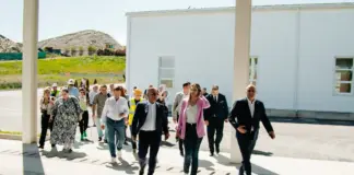 Group of professionally dressed people walking outside in front of a white industrial building on a sunny day, heading toward the camera.