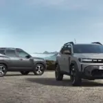 Preturile DACIA Duster 2027 essential | iDevice.ro Two silver Dacia Duster SUVs parked on a rocky mountain pass with cliffs and distant peaks in the background.