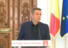 Situatie de Criza in Romania, Sorin Grindeanu cere Masuri Urgente Man in a dark suit speaks at a podium with a PSD banner, flanked by the Romanian and European flags, against ornate wooden doors.