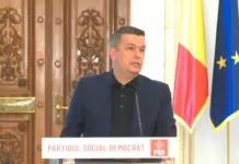 Situatie de Criza in Romania, Sorin Grindeanu cere Masuri Urgente Man in a dark suit speaks at a podium with a PSD banner, flanked by the Romanian and European flags, against ornate wooden doors.