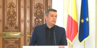 Man in a dark suit speaks at a podium with a PSD banner, flanked by the Romanian and European flags, against ornate wooden doors.