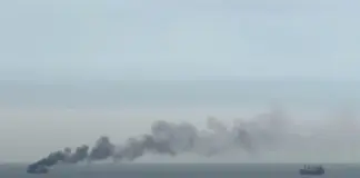 Large ship at sea emits a thick plume of dark smoke to the left, with another cargo vessel visible on the right over calm water.