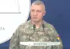 Military officer in camouflage speaking at a podium during a briefing, with a blue backdrop and 'ROMÂNIA' visible on the sign.