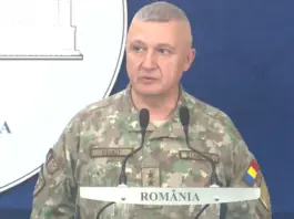 Military officer in camouflage speaking at a podium during a briefing, with a blue backdrop and 'ROMÂNIA' visible on the sign.