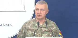 Military officer in camouflage speaking at a podium during a briefing, with a blue backdrop and 'ROMÂNIA' visible on the sign.