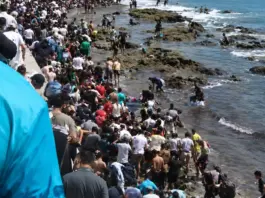 Mass of people crowding a rocky coastline beside the ocean, with some entering the water and others on rocks or a seawall.4a1