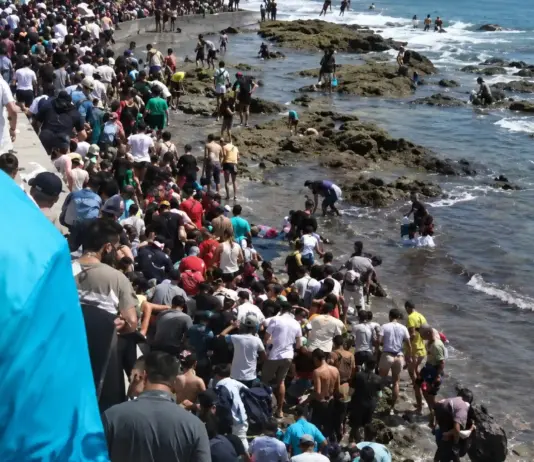 Mass of people crowding a rocky coastline beside the ocean, with some entering the water and others on rocks or a seawall.4a1