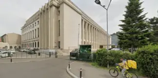 Beige neoclassical building with tall columns beside a street; cyclist with yellow delivery bag rides past.