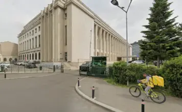 Beige neoclassical building with tall columns beside a street; cyclist with yellow delivery bag rides past.