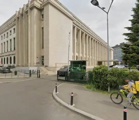Beige neoclassical building with tall columns beside a street; cyclist with yellow delivery bag rides past.