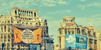 Historic city street with ornate old buildings, blue sky, and large orange banner in the foreground displaying Romanian text.
