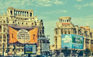 AVERTIZARI Cod ROSU si PORTOCALIU Meteo de ULTIM MOMENT Emise de ANM pentru Romania pe 3 August 2026 Historic city street with ornate old buildings, blue sky, and large orange banner in the foreground displaying Romanian text.