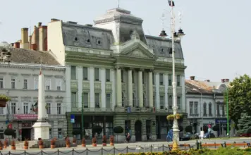 Avertizarile Meteo ANM cu Vremea EXTREMA care Afecteaza mai Multe Regiuni din Romania din 7 August 2026 Historic European-style government building with green facade and ornate pediment, facing a sunny square with lampposts and potted plants.