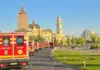 Line of red emergency ambulances (Pompierii/SMURD) driving along a city street past historic yellow stone buildings under a clear blue sky, with green grass and trees on the right.