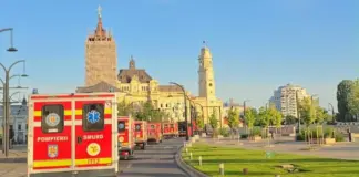 Line of red emergency ambulances (Pompierii/SMURD) driving along a city street past historic yellow stone buildings under a clear blue sky, with green grass and trees on the right.