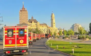 Line of red emergency ambulances (Pompierii/SMURD) driving along a city street past historic yellow stone buildings under a clear blue sky, with green grass and trees on the right.