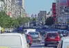 Busy city avenue filled with cars, taxis, and a bus in heavy traffic between green trees on the left and tall buildings with billboards on the right.