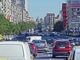 Busy city avenue filled with cars, taxis, and a bus in heavy traffic between green trees on the left and tall buildings with billboards on the right.