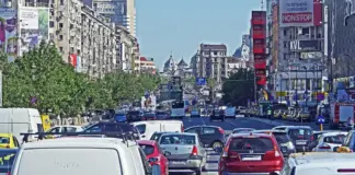 Busy city avenue filled with cars, taxis, and a bus in heavy traffic between green trees on the left and tall buildings with billboards on the right.