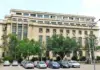 Facade of a large beige office building with rows of windows, green trees, and a parking lot in the foreground.