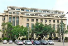 Facade of a large beige office building with rows of windows, green trees, and a parking lot in the foreground.